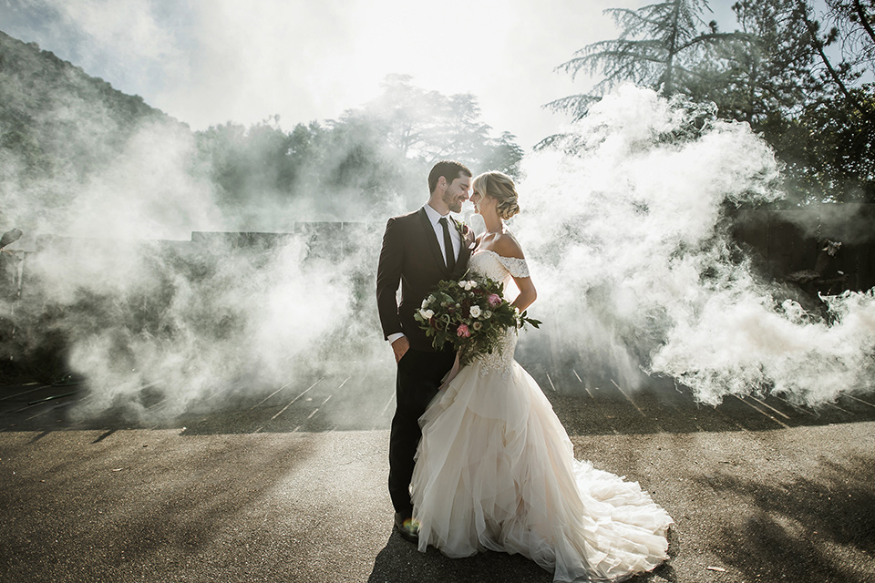 saddle-peak-shoot-bride-and-groom-with-smoke-behind-them-bride-in-an-off-the-shoulder-gown-with-lace-detailing-and-hair-up-in-a-bun-groom-in-a-burgundy-tuxedo-with-a-long-tie