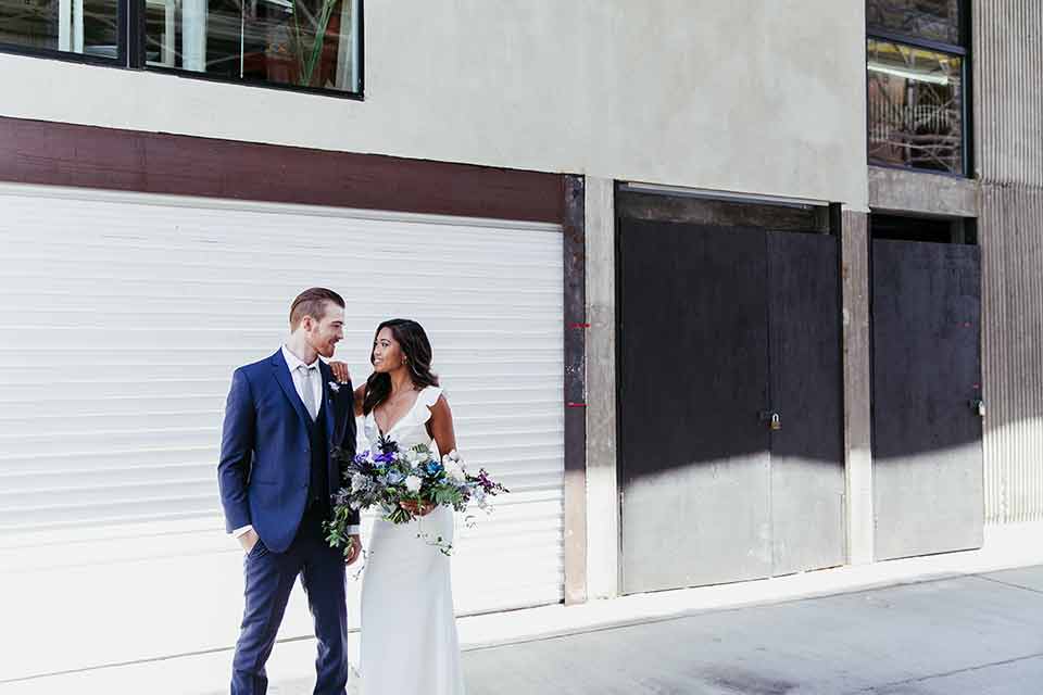 Sandbox-styled-shoot-bride-and-groom-looking-at-each-other-outside-near-white-wall-bride-with-arm-on-groom-shoulder-bride-in-white-gown-with-flutter-sleeves-and-deep-v-neckline-groom-in-blue-suit-with-grey-tie-and-brown-shoes