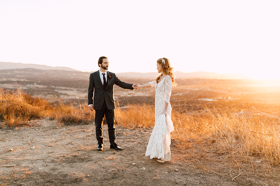 Temecula-wedding-at-wolf-feather-honey-farm-bride-form-fitting-lace-gown-with-sleeves-and-open-back-design-and-groom-charcoal-grey-notch-lapel-suit-with-white-dress-shirt-and-long-burgundy-matte-tie-dancing-holding-hands