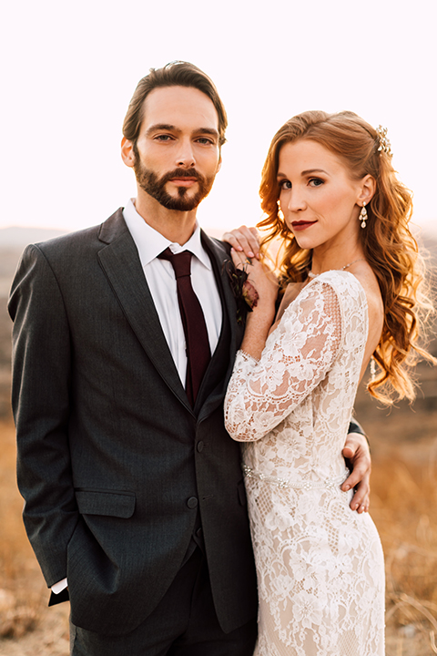 Temecula-wedding-at-wolf-feather-honey-farm-bride-form-fitting-lace-gown-with-sleeves-and-open-back-design-and-groom-charcoal-grey-notch-lapel-suit-with-white-dress-shirt-and-long-burgundy-matte-tie-hugging