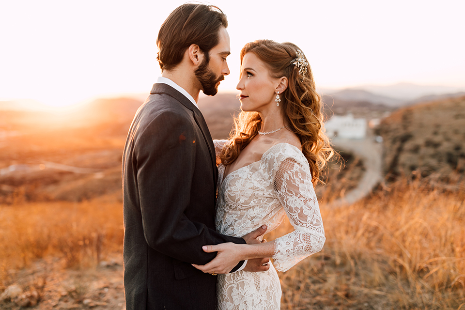 Temecula-wedding-at-wolf-feather-honey-farm-bride-form-fitting-lace-gown-with-sleeves-and-open-back-design-and-groom-charcoal-grey-notch-lapel-suit-with-white-dress-shirt-and-long-burgundy-matte-tie-hugging