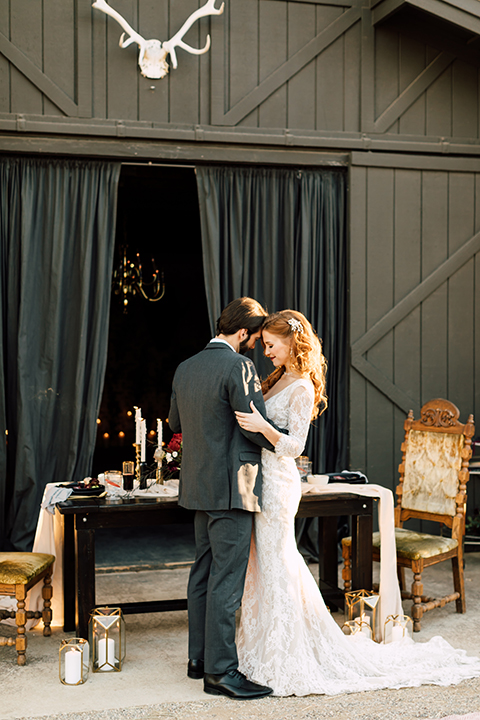 Temecula-wedding-at-wolf-feather-honey-farm-bride-form-fitting-lace-gown-with-sleeves-and-open-back-design-and-groom-charcoal-grey-notch-lapel-suit-with-white-dress-shirt-and-long-burgundy-matte-tie-hugging-standing-by-table