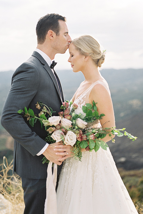 mountain-elopement-shoot-groom-kissing-bride-head-Bride-in-a-tulle-ball-gown-with-an-illusion-bodice-and-lace-detailing-groom-at-sweetheart-table-groom-in-a-grey-tuxedo-with-a-black-bowtie