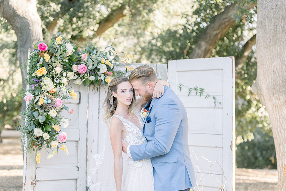 alice-in-wonderland-shoot-bride-and-groom-at-ceremony-bride-in-a-white-gown-with-a-flowing-skirt-and-low-back-detail-groom-in-a-light-blue-coat-with-dark-blue-pants-and-vest-and-a-floral-long-tie