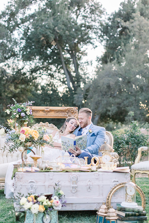 alice-in-wonderland-shoot-bride-and-groom-reading-bride-in-a-white-gown-with-lace-detailing-and-a-flowing-skirt-groom-in-a-light-blue-coat-with-dark-blue-pants-and-a-floral-long-tie