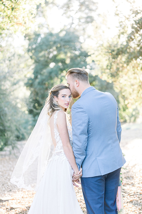 alice-in-wonderland-shoot-bride-and-groom-walking-away-bride-in-a-white-gown-with-a-flowing-white-gown-and-a-deep-back-detail-and-the-groom-in-a-light-blue-suit-with-dark-blue-pants-and-white-floral-long-tie