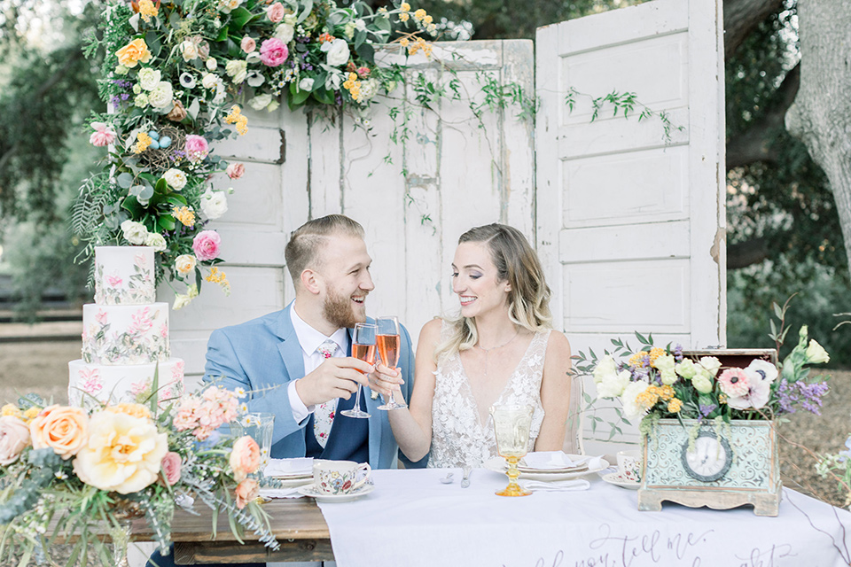 alice-in-wonderland-shoot-couple-at-sweetheart-table-bride-in-a-white-gown-with-a-flowing-skirt-and-low-back-detail-groom-in-a-light-blue-coat-with-dark-blue-pants-and-vest-and-a-floral-long-tie