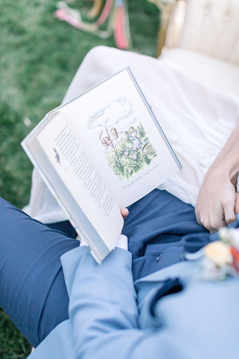 alice-in-wonderland-shoot-couple-reading-a-book-bride-in-a-white-gown-with-a-flowing-white-gown-and-a-deep-back-detail-and-the-groom-in-a-light-blue-suit-with-dark-blue-pants-and-white-floral-long-tie