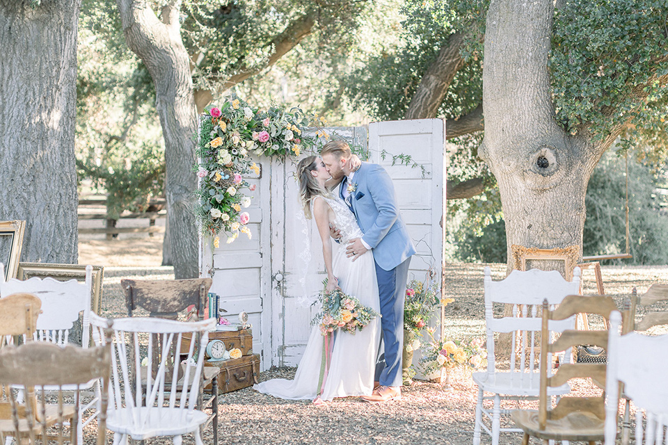 alice-in-wonderland-shoot-first-kiss-bride-in-a-white-gown-with-a-flowing-skirt-and-low-back-detail-groom-in-a-light-blue-coat-with-dark-blue-pants-and-vest-and-a-floral-long-tie