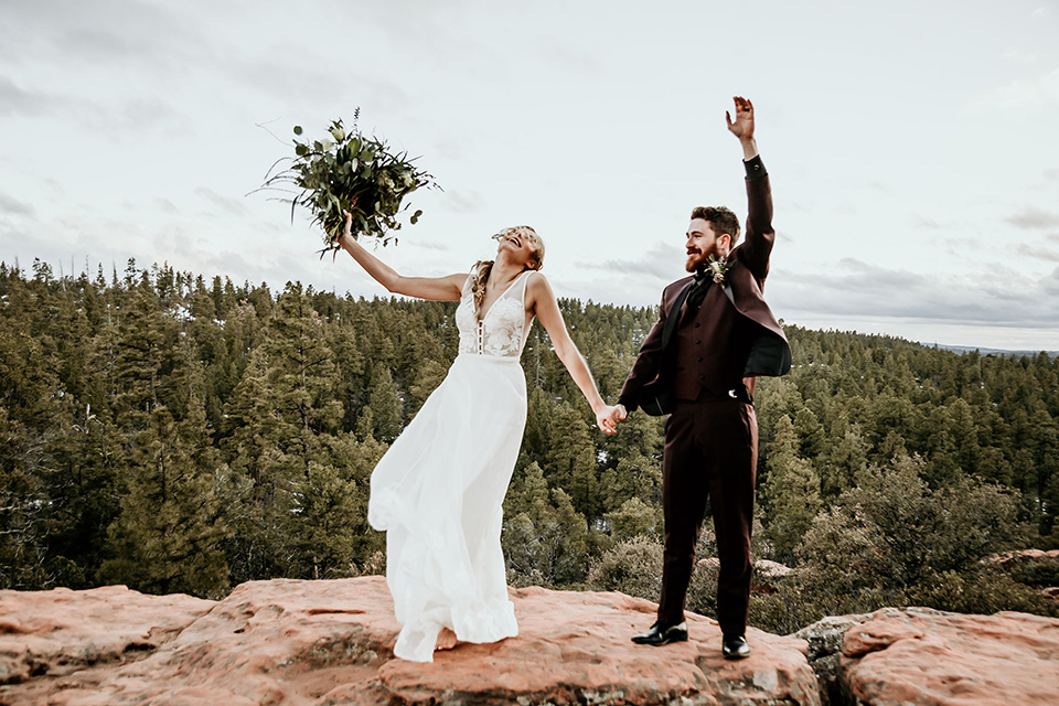 Arizona-Elopement-Shoot-bride-and-groom-celebrating-the-bride-in-a-bohemian-white-gown-with-a-plunging-back-and-lace-detailing-the-groom-in-a-burgundy-shawl-lapel-tuxedo-with-a-black-shirt-and-bow-tie