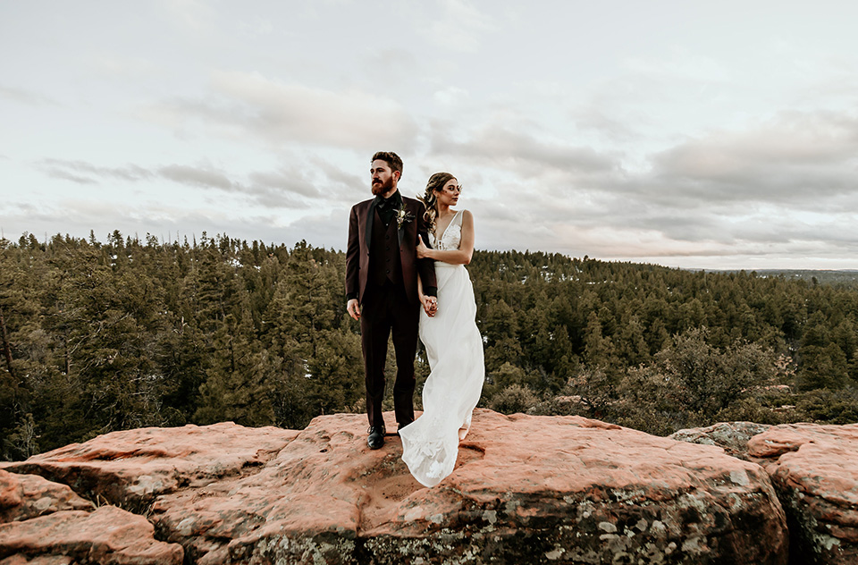 Arizona-Elopement-Shoot-bride-and-groom-looking-to-the-distance-the-bride-in-a-bohemian-white-gown-with-a-plunging-back-and-lace-detailing-the-groom-in-a-burgundy-shawl-lapel-tuxedo-with-a-black-shirt-and-bow-tie