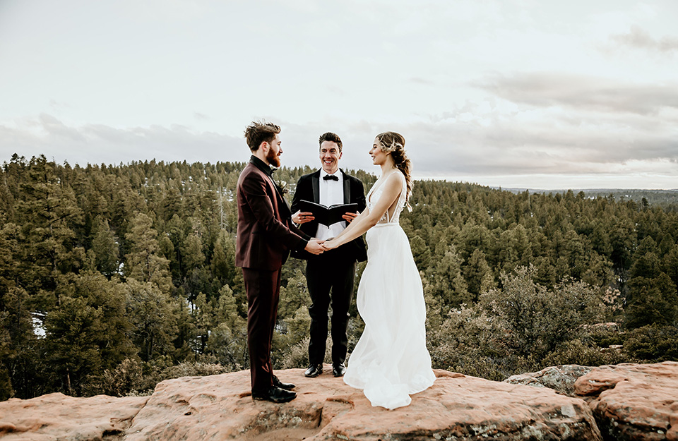 Arizona-Elopement-Shoot-bride-and-groom-with-officiant-on-mountain-the-bride-in-a-bohemian-white-gown-with-a-plunging-back-and-lace-detailing-the-groom-in-a-burgundy-shawl-lapel-tuxedo-with-a-black-shirt-and-bow-tie