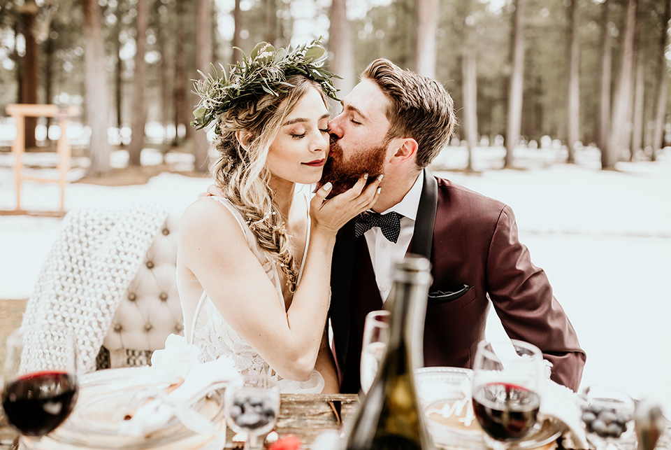 Arizona-Elopement-Shoot-groom-kissing-bride-holding-grooms-face-the-bride-in-a-bohemian-white-gown-with-a-plunging-back-and-lace-detailing-the-groom-in-a-burgundy-shawl-lapel-tuxedo-with-a-black-shirt-and-bow-tie