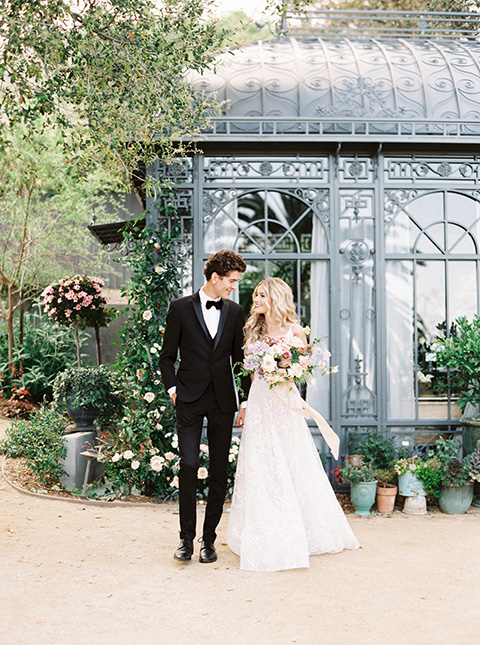 Artist-and-his-muse-shoot-bride-and--groom-walking-by-glass-house-the-bride-in-a-white-ball-gown-with-an-illusion-neckline-and-laace-detailing-and-the-groom-is-in-a-black-tuxedo-with-a-black-bow-tie