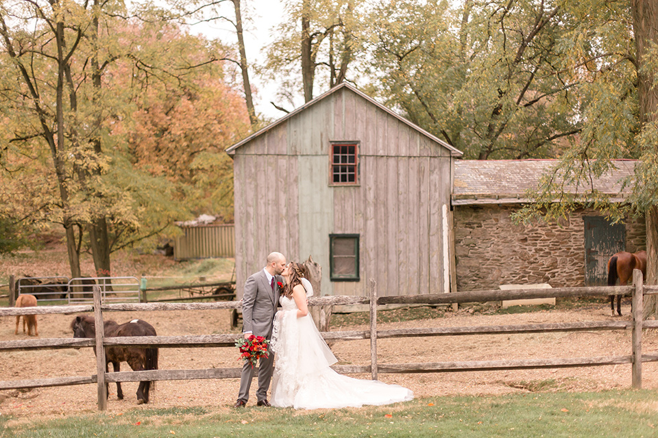 bride in an ivory lace gown with a long veil and train with the groom in a light brown suit with a red long tie and brown shoes, at ranch wedding venue by mini ponies