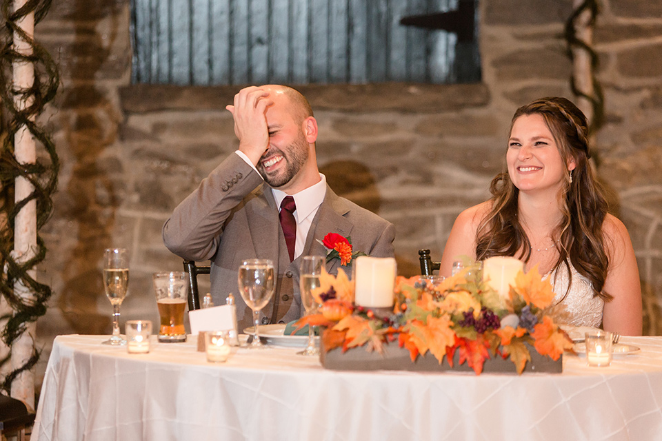 bride in an ivory lace gown with a long veil and train with the groom in a light brown suit with a red long tie and brown shoes, sitting at their sweetheart table