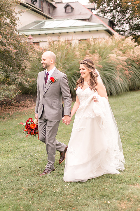  bride in an ivory lace gown with a long veil and train with the groom in a light brown suit with a red long tie and brown shoes, walking at the venue