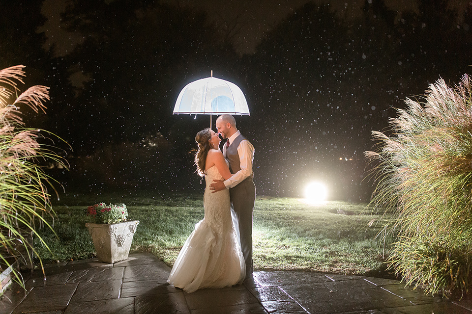 bride in an ivory lace gown with a long veil and train with the groom in a light brown suit with a red long tie and brown shoes, husband and wife with umbrella at night in the rain