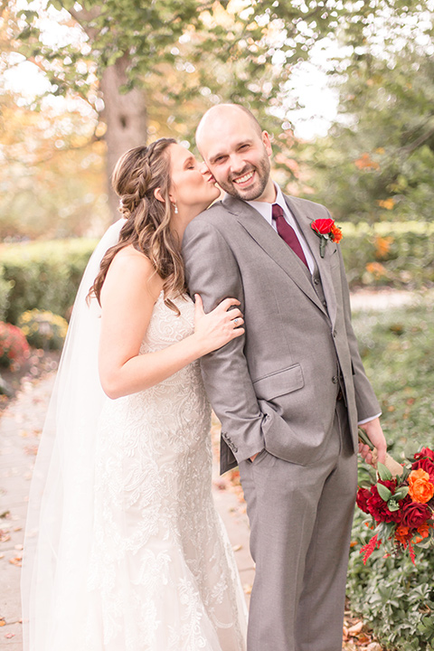 bride in an ivory lace gown with a long veil and train with the groom in a light brown suit with a red long tie and brown shoes, walking at the venue