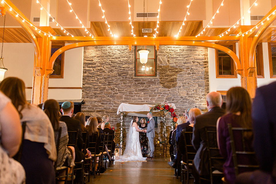 bride in an ivory lace gown with a long veil and train with the groom in a light brown suit with a red long tie and brown shoes, ceremony