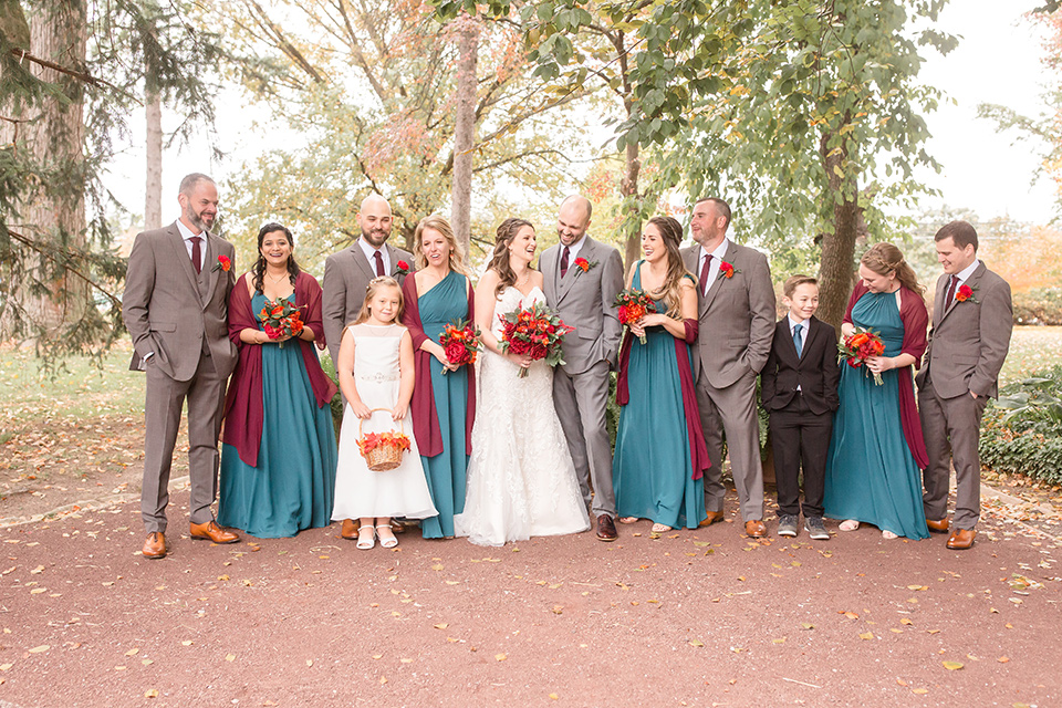 groom and groomsmen in brown suits with red ties and brown shoes with the bride in an ivory lace gown with her bridesmaids in teal colored gowns with red shawls