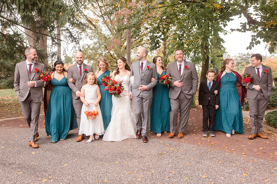 groom and groomsmen in brown suits with red ties and brown shoes with the bride in an ivory lace gown with her bridesmaids in teal colored gowns with red shawls