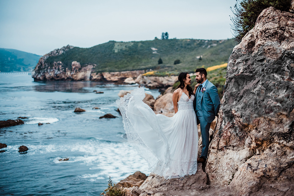 avila-beach-elopement-shoot-bide-and-groom-on-rockside-cliff-bride-in-a-flowing-white-gown-with-a-soft-tulle-overlay-the-groom-wore-a-light-blue-suit-with-a-navy-long-tie