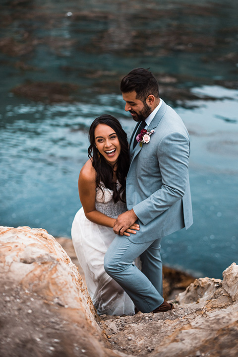 avila-beach-elopement-shoot-bride-laughing-groom-golding-her-steady-bride-in-a-soft-flowing-gown-with-a-strapless-neckline-and-the-groom-in-a-light-blue-suit-with-a-navy-long-tie