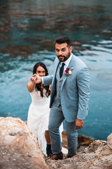 avila-beach-elopement-shoot-groom-helping-bride-up-groom-wearing-a-light-blue-suit-with-a-navy-long-tie-and-bride-in-a-strapless-tulle-flowing-gown