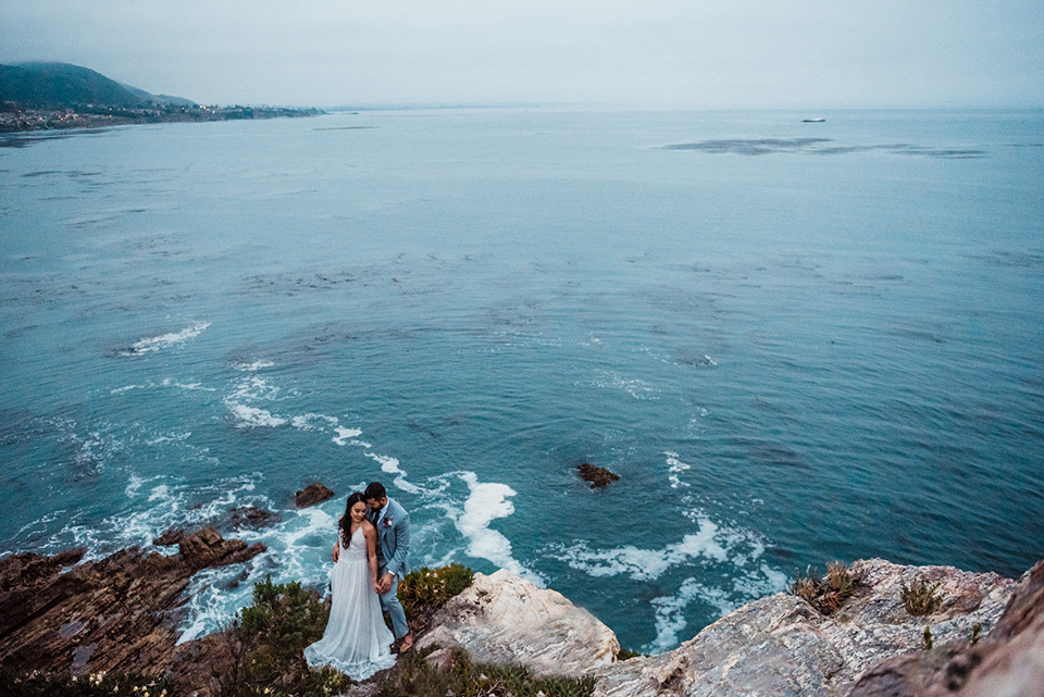 avila-beach-elopement-shoot-long-shot-of-ocean-and-couple-bride-in-a-flowing-white-gown-with-a-soft-tulle-overlay-the-groom-wore-a-light-blue-suit-with-a-navy-long-tie