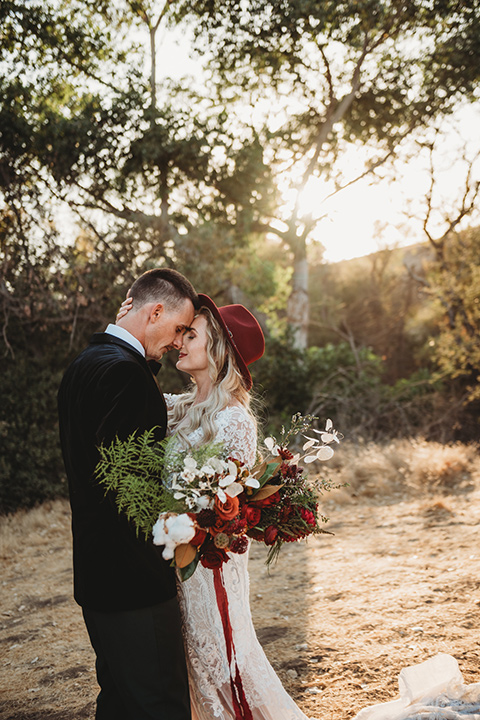 owl-creek-farms-bide-and-groom-touching-heads-owl-creek-farms-bide-and-groom-touching-heads-bride-wearing-a-boho-style-dress-with-long-sleeves-and-a-high-neckline-with-a-burgundy-wide-brimmed-hat-groom-wearing-a-velvet-tuxedo-with-grey-pants