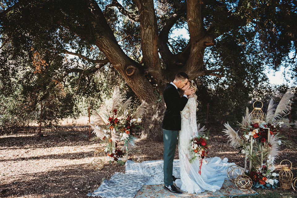 owl-creek-farms-bride-and-groom-kissing-under-tree-bride-wearing-a-boho-lace-long-sleeved-gown-with-a-high-neckline-groom-wears-a-black-velvet-coat-with-grey-pants-and-a-black-bow-tie