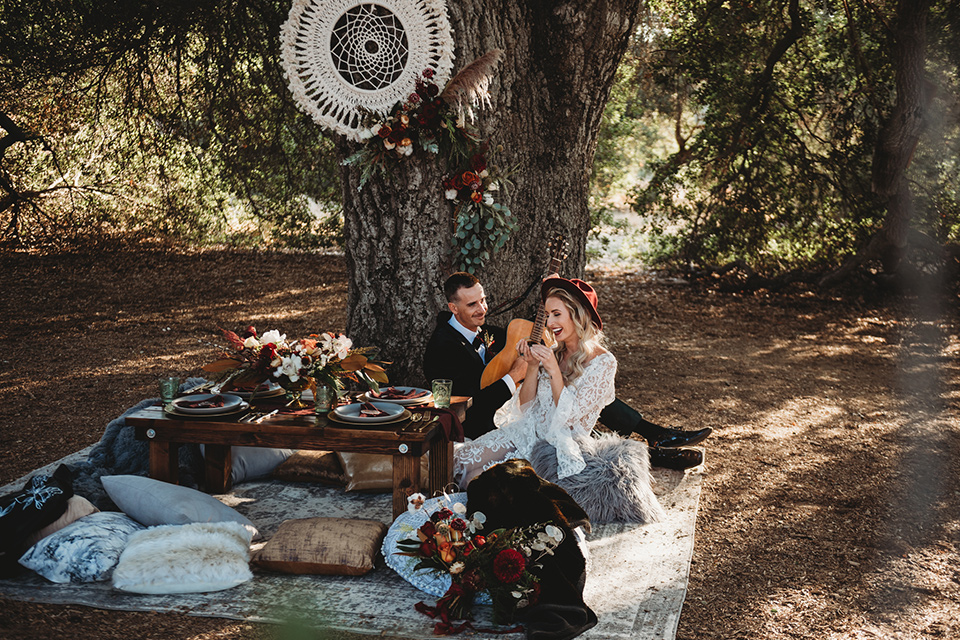 owl-creek-farms-bride-and-groom-sitting-with-instruments-bride-wearing-a-boho-lace-long-sleeved-gown-with-a-high-neckline-groom-wears-a-black-velvet-coat-with-grey-pants-and-a-black-bow-tie