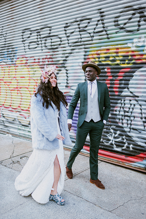 Brooklyn-Shoot-bride-and-groom-looking-at-eachother-walking-by-graffitti-bride-in-a-bohemian-style-gown-and-groom-in-a-green-suit-with-brown-shoes-and-bolo-tie