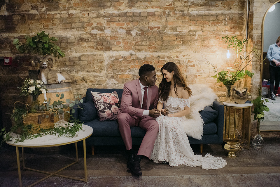 Brooklyn-Shoot-bride-and-groom-sitting-on-couch-groom-in-a-rose-pink-suit-with-brown-shoes-the-bride-in-a-lace-boho-style-gown