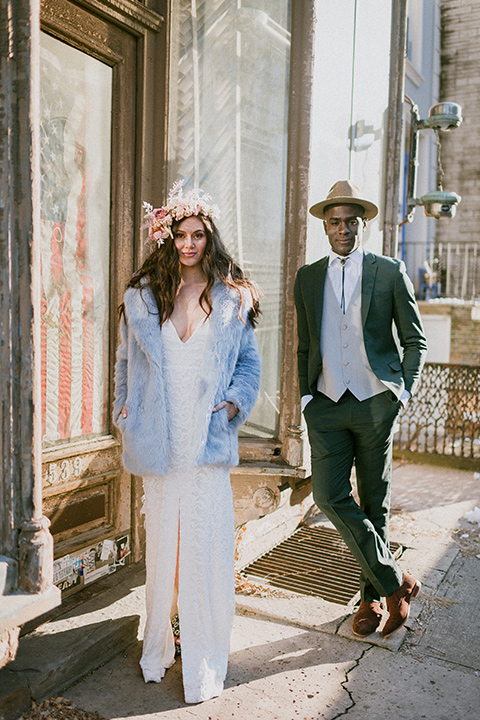 Brooklyn-Shoot-bride-and-groom-standing-against-the-wall-outside-bride-in-a-lace-boho-gown-with-an-off-the-shoulder-detail-and-a-blue-fur-coat-groom-in-a-deep-green-suit-with-brown-shoes-and-a-bolo-tie