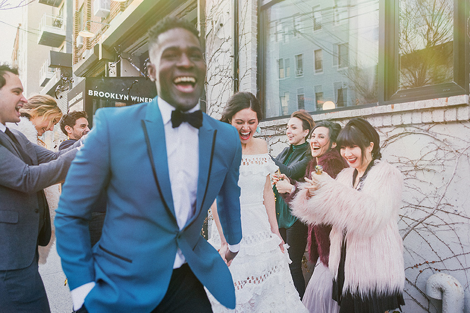 Brooklyn-Shoot-bride-and-groom-walking-close-up-laughing-groom-in-a-teal-coat-with-black-pants-and-a-black-bow-tie-the-bride-wears-a-bohemian-style-gown-with-flutter-sleeves-and-loose-hair