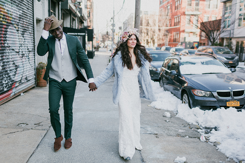 Brooklyn-Shoot-bride-and-groom-walking-outside-groom-in-a-green-suit-with-a-bolo-tie-the-bride-wears-a-bohemian-style-gown-with-flutter-sleeves-and-loose-hair-and-a-blue-faux-fur-coat
