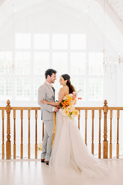 bride and groom close standing in loft, bride in a full tulle ballgown with a strapless design and the groom in a light grey suit with pink bow tie