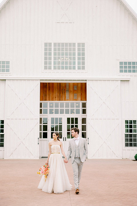 bride and groom walking outside venue, bride in a full tulle ballgown with a strapless design and the groom in a light grey suit with pink bow tie