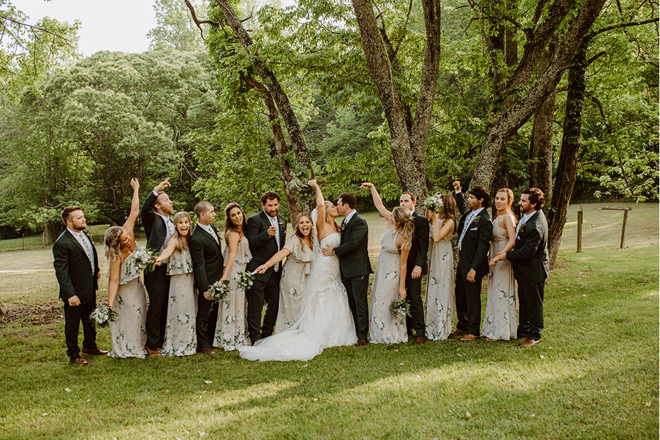 Horse-Range-Vista-bridal-party-hands-in-the-air-bridesmaids-in-sage-dresses-with-green-and-white-flowers-on-it-groomsmen-in-green-suits-with-floral-ties-bride-in-a-lace-mermaid-gown-with-a-long-veil-groom-in-a-green-suit-with-a-floral-tie