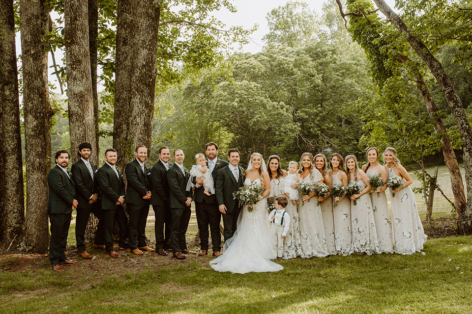 Horse-Range-Vista-bridal-party-standing-bridesmaids-in-sage-dresses-with-green-and-white-flowers-on-it-groomsmen-in-green-suits-with-floral-ties-bride-in-a-lace-mermaid-gown-with-a-long-veil-groom-in-a-green-suit-with-a-floral-tie