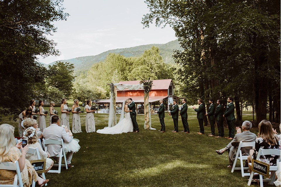 Horse-Range-Vista-ceremony-bridesmaids-in-sage-dresses-with-green-and-white-flowers-on-it-groomsmen-in-green-suits-with-floral-ties-bride-in-a-lace-mermaid-gown-with-a-long-veil-groom-in-a-green-suit-with-a-floral-tie