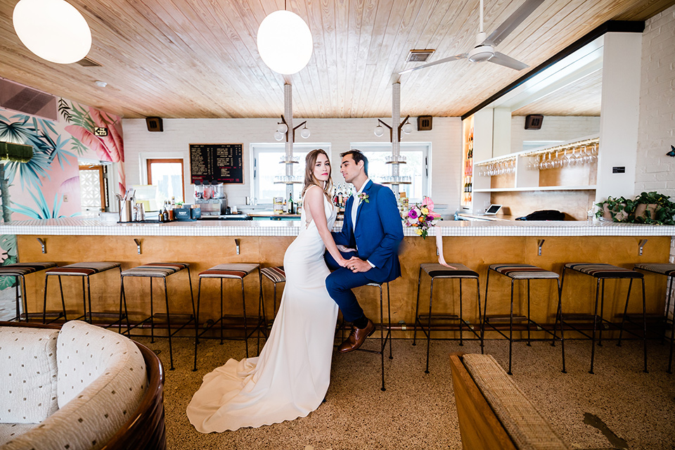 bride and groom at the bar at the drifter hotel, bride is in a formfitting gown with straps and her hair down in a loose style while the groom is in a cobalt blue suit with brown shoes and his shirt undone