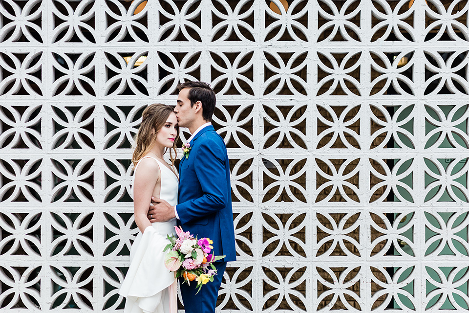 bride and groom outside the venue next to a cinder block wall, bride is in a formfitting gown with straps and her hair down in a loose style while the groom is in a cobalt blue suit with brown shoes and his shirt undone