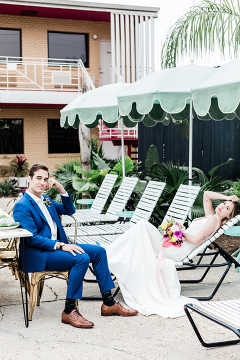 bride and groom on the poolside furniture, bride is in a formfitting gown with straps and her hair down in a loose style, while the groom is in a cobalt blue suit with brown shoes and his shirt undone