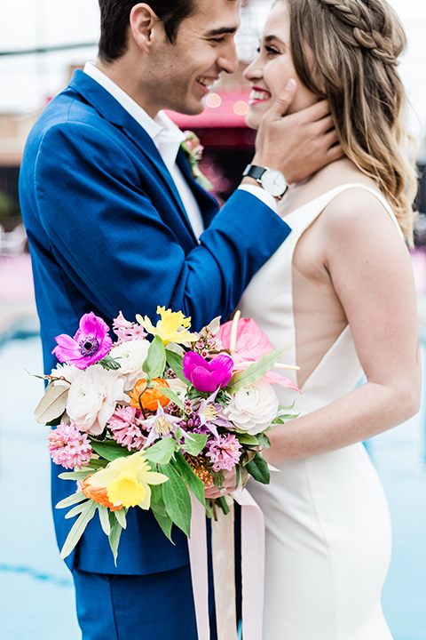 bride and groom close up by the pool, bride is in a formfitting gown with straps and her hair down in a loose style and holding a bright colored bouquet of flowers, while the groom is in a cobalt blue suit with brown shoes and his shirt undone