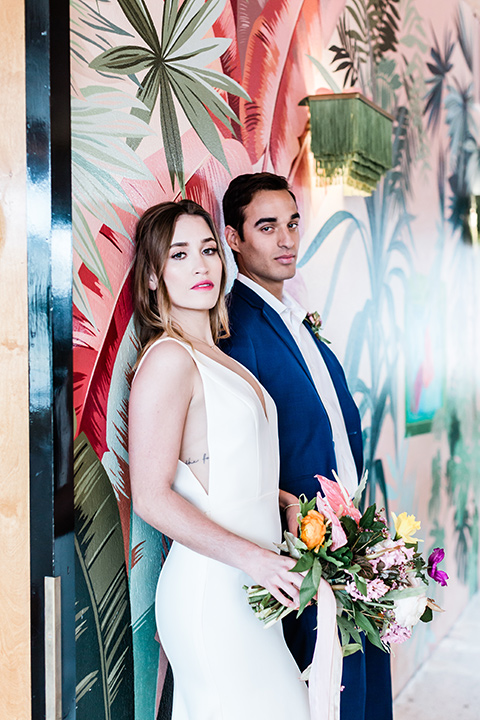 bride and groom standing by a mural, bride is in a formfitting gown with straps and her hair down in a loose style and holding a bright colored bouquet of flowers, while the groom is in a cobalt blue suit with brown shoes and his shirt undone