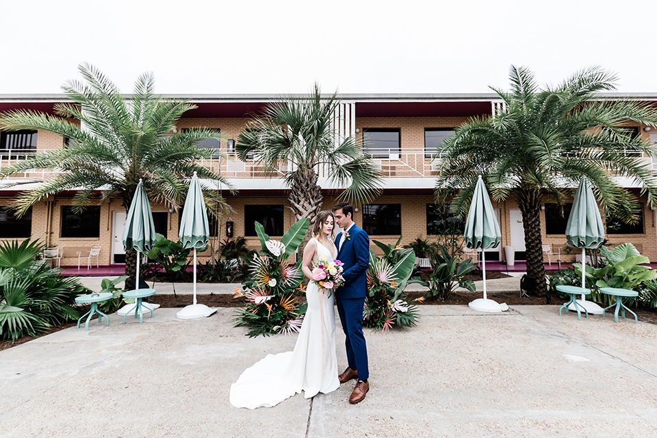 bride and groom standing close outside the hotel, bride is in a formfitting gown with straps and her hair down in a loose style while the groom is in a cobalt blue suit with brown shoes and his shirt undone