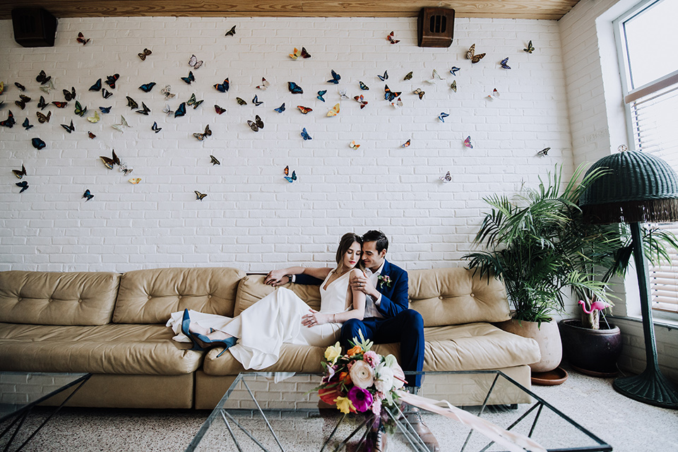 bride and groom on the couch with a butterfly art piece above them, bride is in a formfitting gown with straps and her hair down in a loose style while the groom is in a cobalt blue suit with brown shoes and his shirt undone